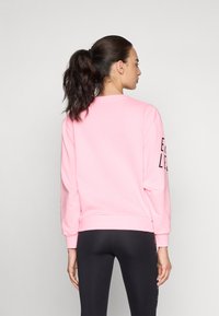 Femme aux cheveux bruns en queue de cheval portant un sweat-shirt rose et un legging noir, de dos devant un fond gris uni.