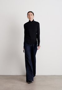 Zwarte turtleneck, slim fit, in combinatie met brede, donkere spijkerbroeken. Model houdt een klein metallic voorwerp in haar hand. Eenvoudige achtergrond.