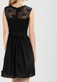 Robe noire avec un corsage en dentelle à encolure arrondie, sans manches, et une jupe en satin lisse et plissée s'étendant jusqu'aux genoux.
