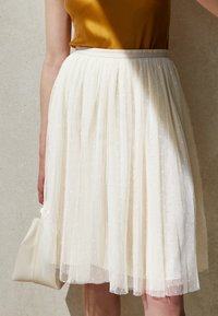 Nainen, jolla on päällään valkoinen polvipituinen tüllihame, jossa on pieniä ristikuviota, ja sinapinkeltainen satiinitoppi, pitää kermansävyistä käsilaukkua kyljellään.