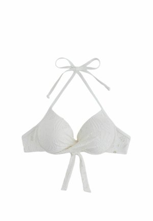 Kvinde med langt mørkt hår iført en hvid tekstureret bikinitop, der står foran en solbeskinnet klippebaggrund.