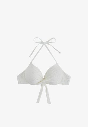 Kvinde med langt mørkt hår iført en hvid tekstureret bikinitop, der står foran en solbeskinnet klippebaggrund.