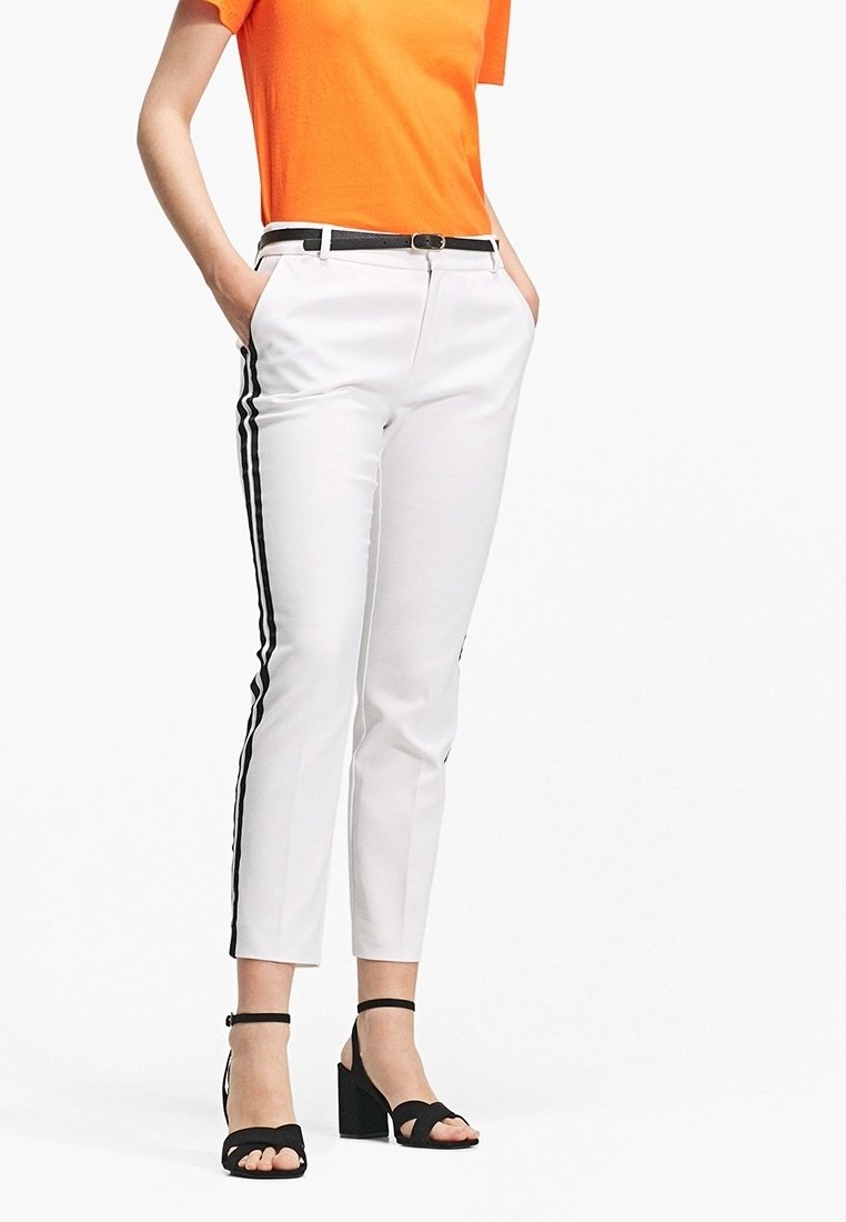 Osoba ubrana w białe spodnie o krótkiej długości z czarnymi paskami po bokach, czarny pasek, pomarańczową bluzkę i czarne sandały na słupku, stoi z rękami w kieszeniach.