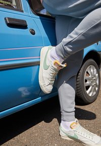 Valkoiset ja vihreät Nike Blazer -lenkkarit keltaisilla yksityiskohdilla ja teksturoiduilla yksityiskohdilla, yhdistettyinä harmaisiin jogging-housuihin, sinisen auton vieressä.