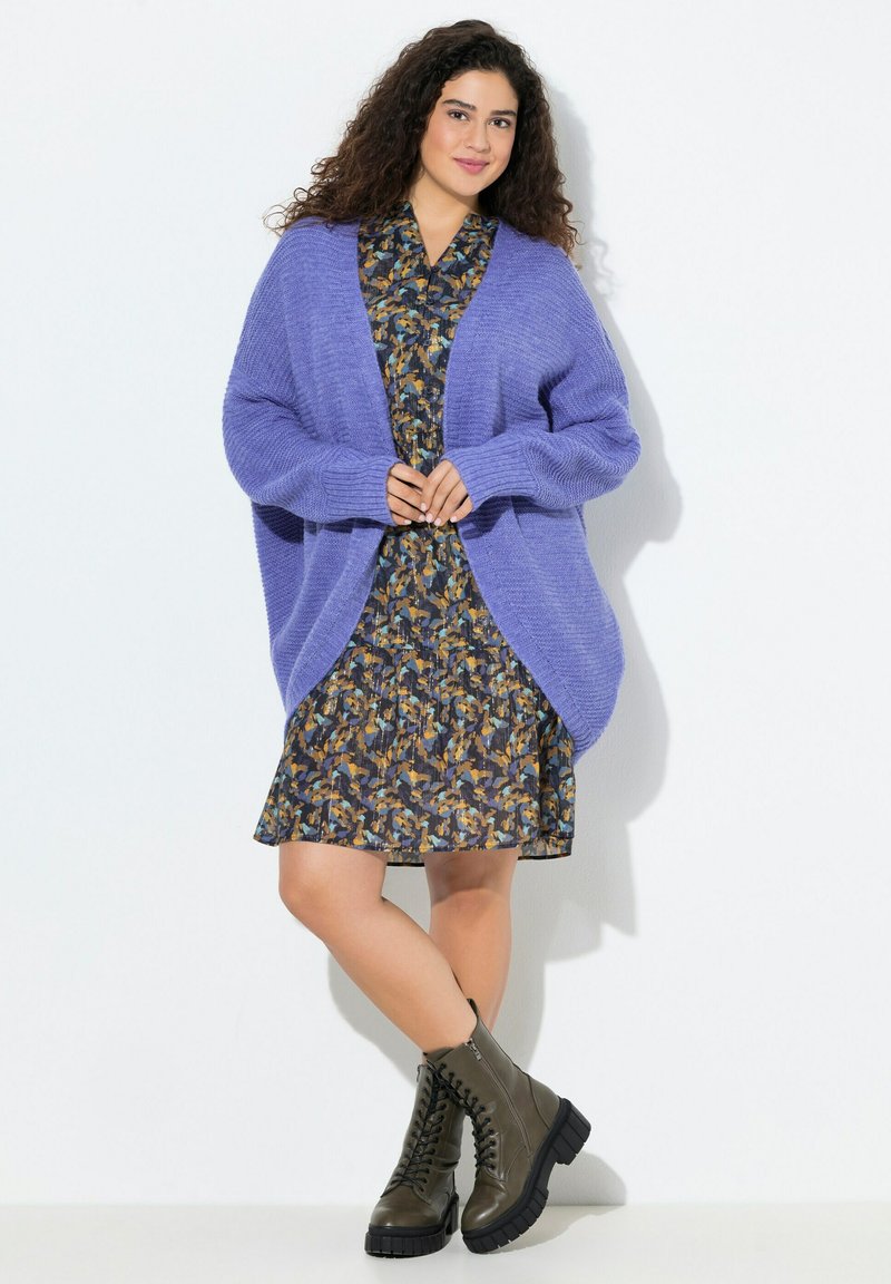 Lila stickad cardigan med neddragna axlar över en blommig klänning i blått, gult och brunt. Matchas med svarta snörskor.