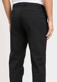 Pantaloni neri con tessuto testurizzato, vestibilità su misura e due tasche posteriori con dettagli di cucitura discreti, indossati con una camicia bianca.