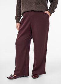 Personne portant un pantalon large bordeaux, un pull marron et des mocassins bordeaux avec boucle et détails à clous, debout avec une main dans la poche.