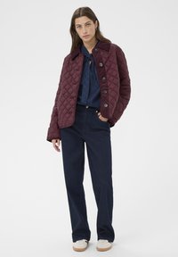 Bordeaux farbener, gesteppter Jacke mit Kragen, großen Knöpfen und lockerer Passform. Kombiniert mit dunkelblauen weit geschnittenen Hosen und weißen Schuhen.