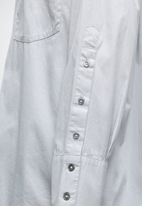 Világosszürke, hosszú ujjú ingujj öt fémből készült gombbal és látható varrással, az anyagon részben megjelenő zsebbel.