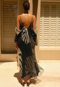 Robe longue dos nu, en tissu transparent avec une base sombre ornée de motifs légers de feuillage. Comprend des bretelles fines et des manches fluides.