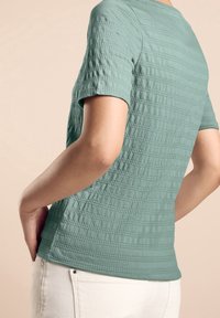 Zielona, teksturowana koszula z krótkim rękawem w kratkę, z okrągłym dekoltem. Połączona z jasnymi spodniami.