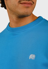 Svetlo modrá mikina s rebrovým okrúhlym výstrihom a malým, vyšitým bielym logom slona na hrudi. Mäkkučká textúra.