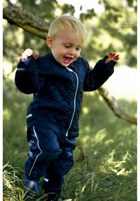 Tumši zilas krāsas pūkaina jaka un bikses komplekts ar priekšējo rāvējslēdzēju, baltām detaļām un ribotām aprocēm, ko nēsā bērns ārā uz zāles.