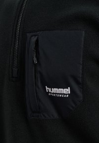Sorte fleece beklædning med en lynlås lomme på brystet i et glat stof. Lommen har en trækkrog, der viser "hummel" i hvid tekst.