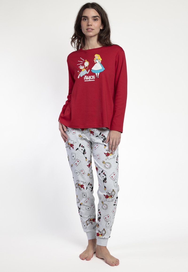 Joven mujer vistiendo una camiseta de manga larga roja con un estampado de Alicia en el País de las Maravillas y pantalones de pijama gris claro que presentan diseños de cartas de juego, relojes y conejos.