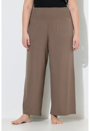 Persoon die losse, hooggetailleerde, bruine wijde broek met elastische tailleband draagt, blootsvoets staand tegen een witte muur.