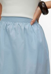 Lyseblå, knælange nederdel med indsamlet taljedetalje. Stoffet virker blødt og glat, suppleret med en enkel, tætsiddende hvid top og mørkt tilbehør.
