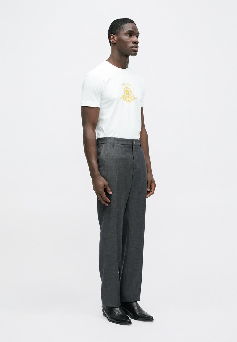 Wit T-shirt met gouden logo, gecombineerd met grijze, op maat gemaakte broek. Zwarte leren schoenen maken de outfit compleet. Simpele, moderne stijl.