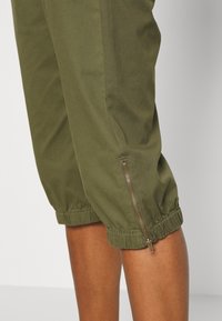 Olivově zelené zkrácené kalhoty s elastickými manžetami a bočním zipem. Tkanina vypadá hladce s mírným leskem.