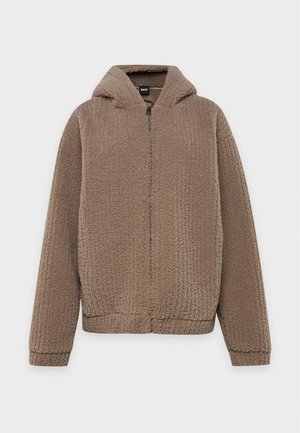 Un homme se tient devant un fond uni, vêtu d’une veste en maille marron à capuche, d’une chemise crème, d’un pantalon à carreaux et de chaussures marron.