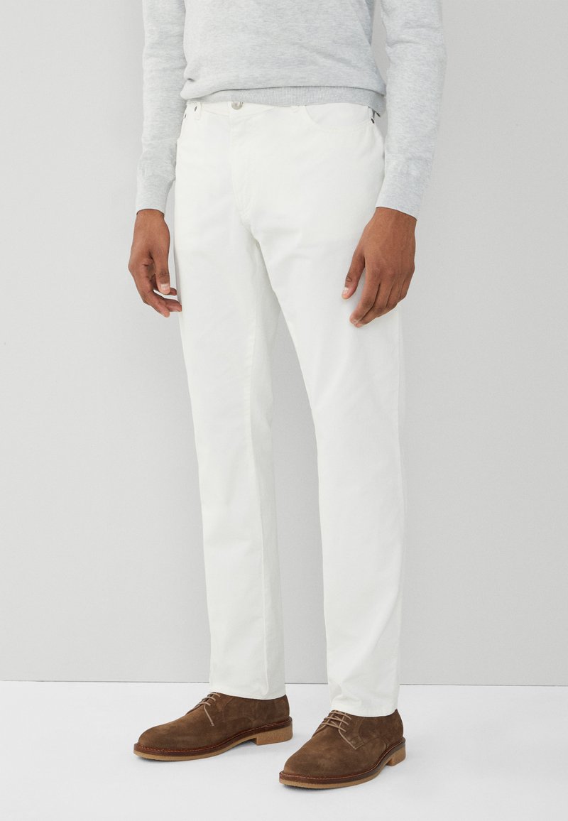 Pantalones de algodón blancos con corte recto, que cuentan con dos bolsillos y cierres metálicos, combinados con zapatos de ante marrones.
