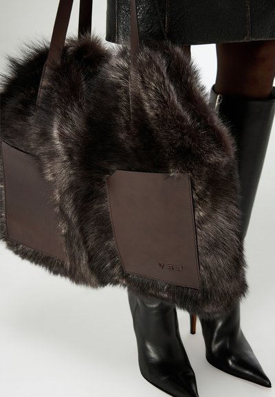 Borsa tote in ecopelliccia marrone con dettagli in pelle, caratterizzata da due tasche rettangolari. Stivali neri con tacco alto parzialmente visibili in primo piano.