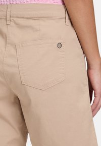 Shorts en coton beige avec une poche arrière à patch avec un bord arrondi et cousu, ainsi qu'un accent de logo en métal. Texture lisse et coupe ajustée.