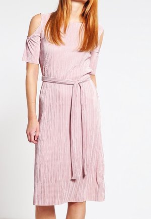 Vrouw in een lichtroze, geribbelde midi-jurk met koude schoudermouwen en een bijpassende riem die om de taille is geknoopt, tegen een witte achtergrond.