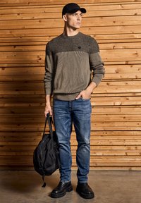 Dunkelolivgrüner Strickpullover mit strukturierter Oberseite, blaue Jeans, schwarze robuste Stiefel und ein schwarzer Rucksack, vor einem Holz-Hintergrund.