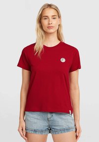 Red cotton t-shirt with a round neckline and short sleeves, featuring a small circular logo on the left chest. Paired with light denim shorts.