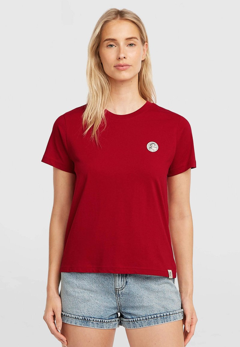 Red cotton t-shirt with a round neckline and short sleeves, featuring a small circular logo on the left chest. Paired with light denim shorts.