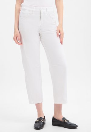 Femme portant un pantalon blanc raccourci et des mocassins noirs slip-on avec une boucle en métal doré, debout sur un fond blanc.