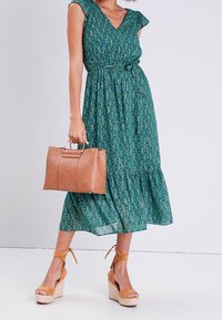 Robe florale verte avec des manches courtes à volants et une taille cintrée, associée à un sac à main en cuir beige et des sandales à plateforme beiges.