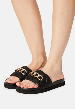 Sorte quiltede tøfler med en tyk sål, der har en guldkæde som accent over overdelen. Glat lædertekstur og åbent tå-design.