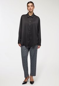 Kobieta stoi w luźnej czarnej satynowej koszuli na guziki, szarych spodniach w kratę i czarnych szpilkach zakończonych na ostro na białym tle.