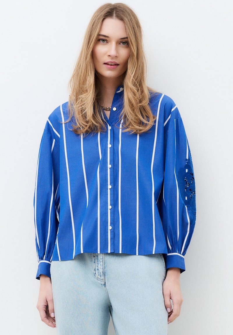 Femme portant une chemise bleue à boutons avec des rayures blanches verticales et des détails brodés aux coudes, associée à un jean bleu clair.