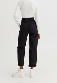 Persoon in zwarte hoge taille paperbag broek, witte long-sleeve top en witte hoge sneakers, staand tegen een effen achtergrond.