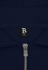 Sweat-shirt à capuche zippé bleu marine avec une fermeture éclair argentée. Présente une étiquette noire avec un label blanc "B". Tissu doux avec une texture lisse.