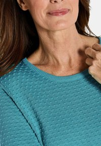 Frau mit langen braunen Haaren, die einen strukturierten türkisfarbenen Pullover trägt, leicht lächelt und eine Hand in der Nähe ihrer Schulter hat, vor einem weißen Hintergrund.
