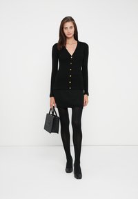 Black ribbed cardigan with a V-neck, gold buttons, paired with a black knit skirt, black tights, and a small handbag. Smooth texture.