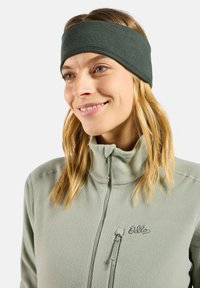 Un bandeau en tissu vert foncé avec une texture lisse, porté avec une veste polaire zippée vert clair dotée d'une petite poche zippée.