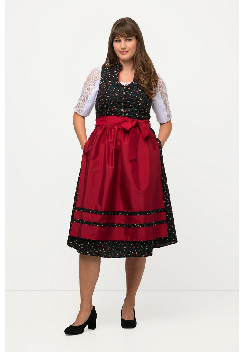 Vêtement traditionnel avec un corsage noir à motifs floraux et un tablier en satin rouge. Présente une taille ceinturée et une jupe à mi-mollet, assortie à des talons noirs.