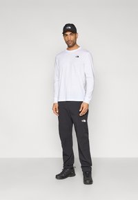 Λευκό πουκάμισο με μακριά μανίκια και μαύρο λογότυπο, συνδυασμένο με μαύρο παντελόνι που φέρει το ίδιο λογότυπο. Το σύνολο ολοκληρώνουν μαύρο καπέλο και μαύρα παπούτσια πεζοπορίας.