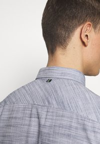 Camisa de cuello en tejido de color azul claro. Patrón texturizado, con un pequeño logotipo en verde en la parte trasera del cuello. Corte corto y ajustado.