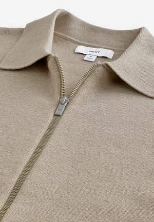 Beige zip-up sweater med et tekstureret strikstof, der har en klassisk krave og metal lynlås. Taget viser størrelse M.