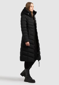 Cappotto imbottito nero con una silhouette lunga, grande cappuccio e dettagli trapuntati. Presenta una zip frontale e tasche laterali; abbinato a stivali neri.