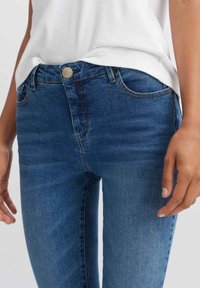 Detailní záběr osoby v modrých denimových džínách středního střihu a bílém tričku zasunutém uvnitř, který ukazuje přední kapsy a detaily knoflíků.