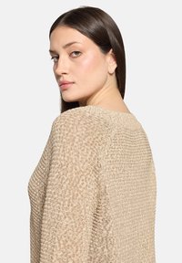Pull beige tricoté avec un motif texturé, un col large et une coupe décontractée, présentant un subtil côtelé le long des coutures et de l'ourlet.