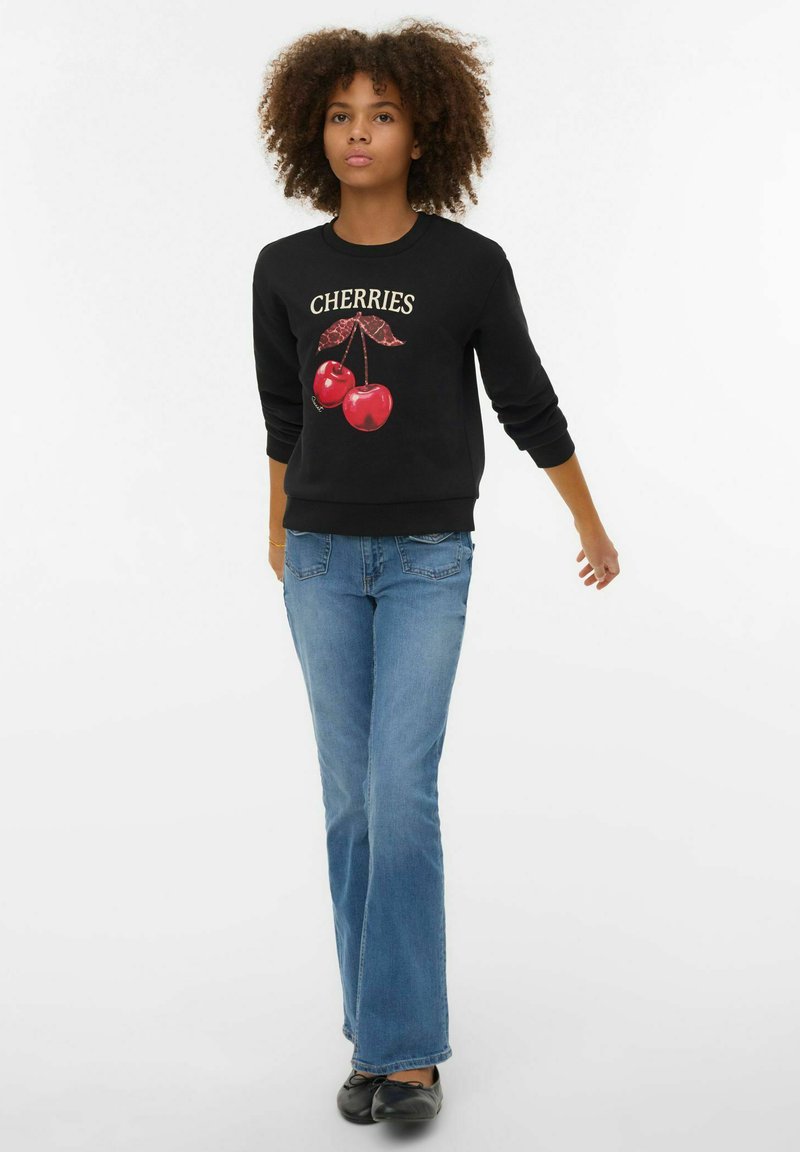 Melna džemperis ar uzrakstu "CHERRIES" un sarkanu ķiršu grafiku. Kombinēts ar gaiši ziliem izpletušiem džinsiem un melniem apaviem. Gluda auduma.