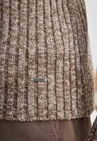 Brauner gestrickter Pullover mit vertikaler Rippenstruktur, ausgestattet mit einem silbernen Logo-Tag. Aus weichem Garn und einem mehrschichtigen Muster in neutralen Farbtönen.